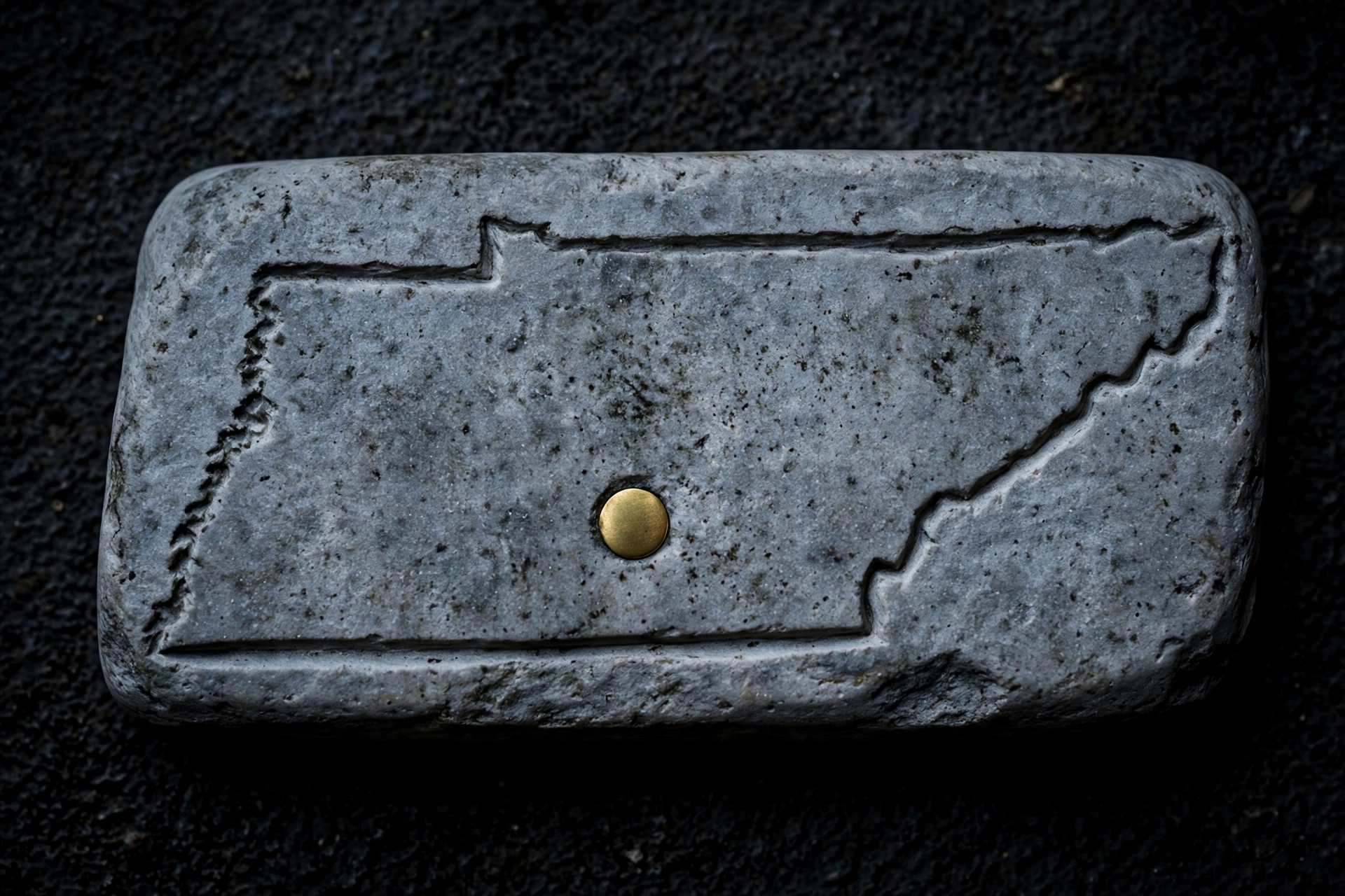 A photograph of a gray rock with an outline of the state of Tennessee carved into it and a brass tack at its center.