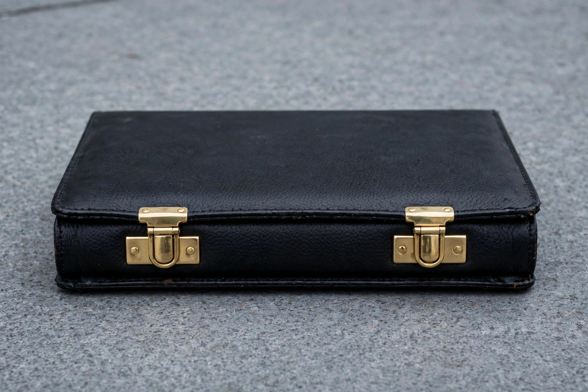 A photograph of a closed black briefcase with gold clasps resting on a grey concrete surface.