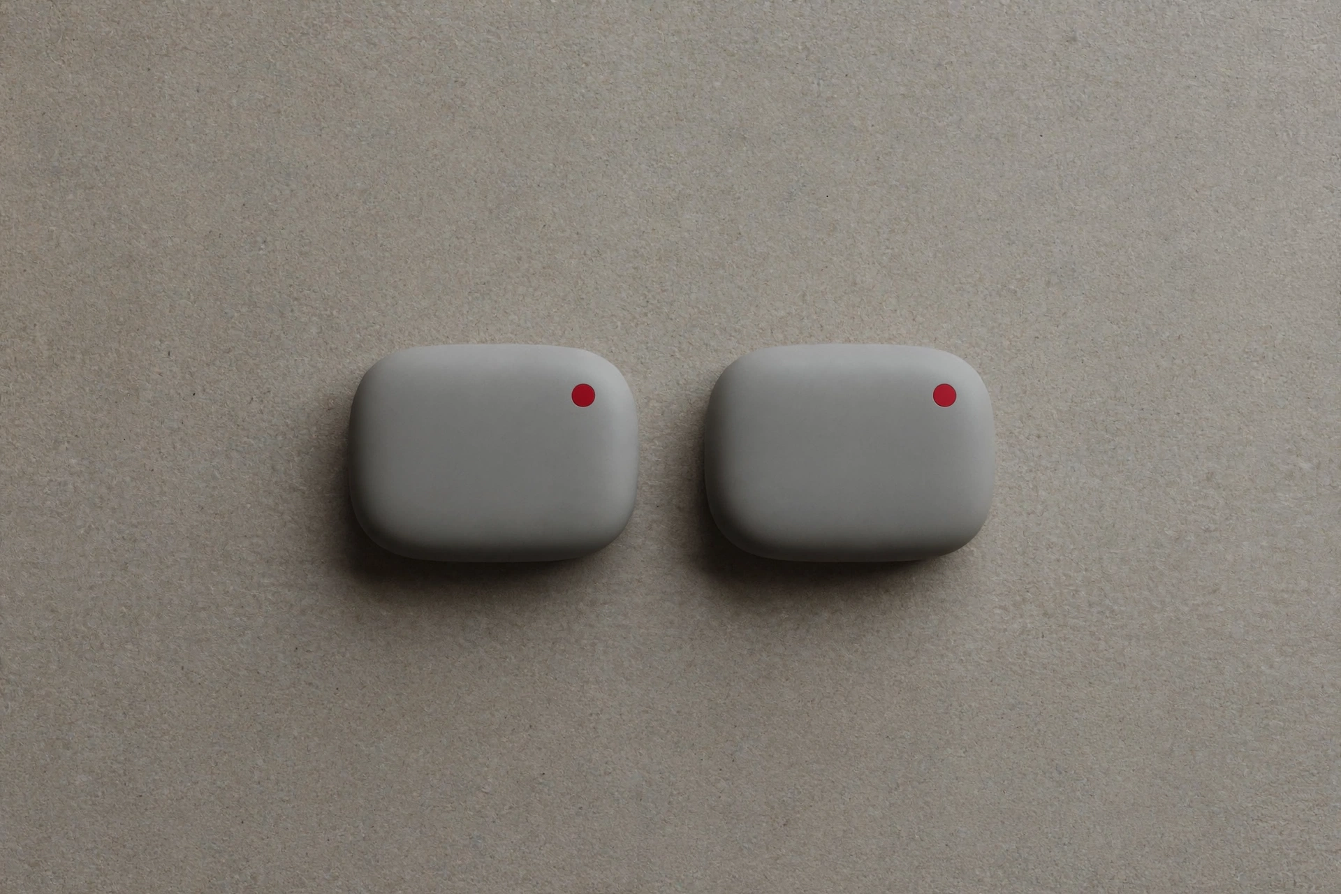 A photograph of two small, light gray, rounded square objects with a red dot on each, placed on a textured gray background.
