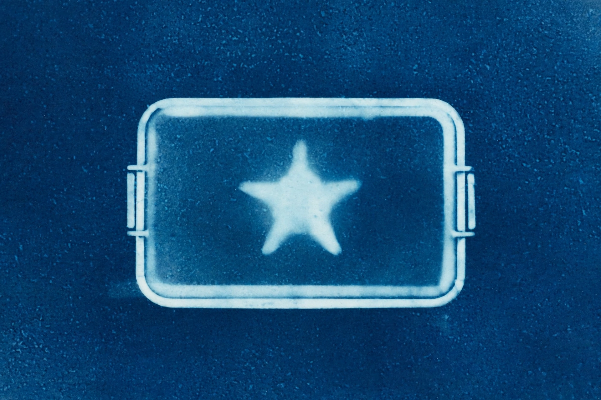 A cyanotype photograph shows a rectangular object with two handles and a star inside on a speckled blue background.
