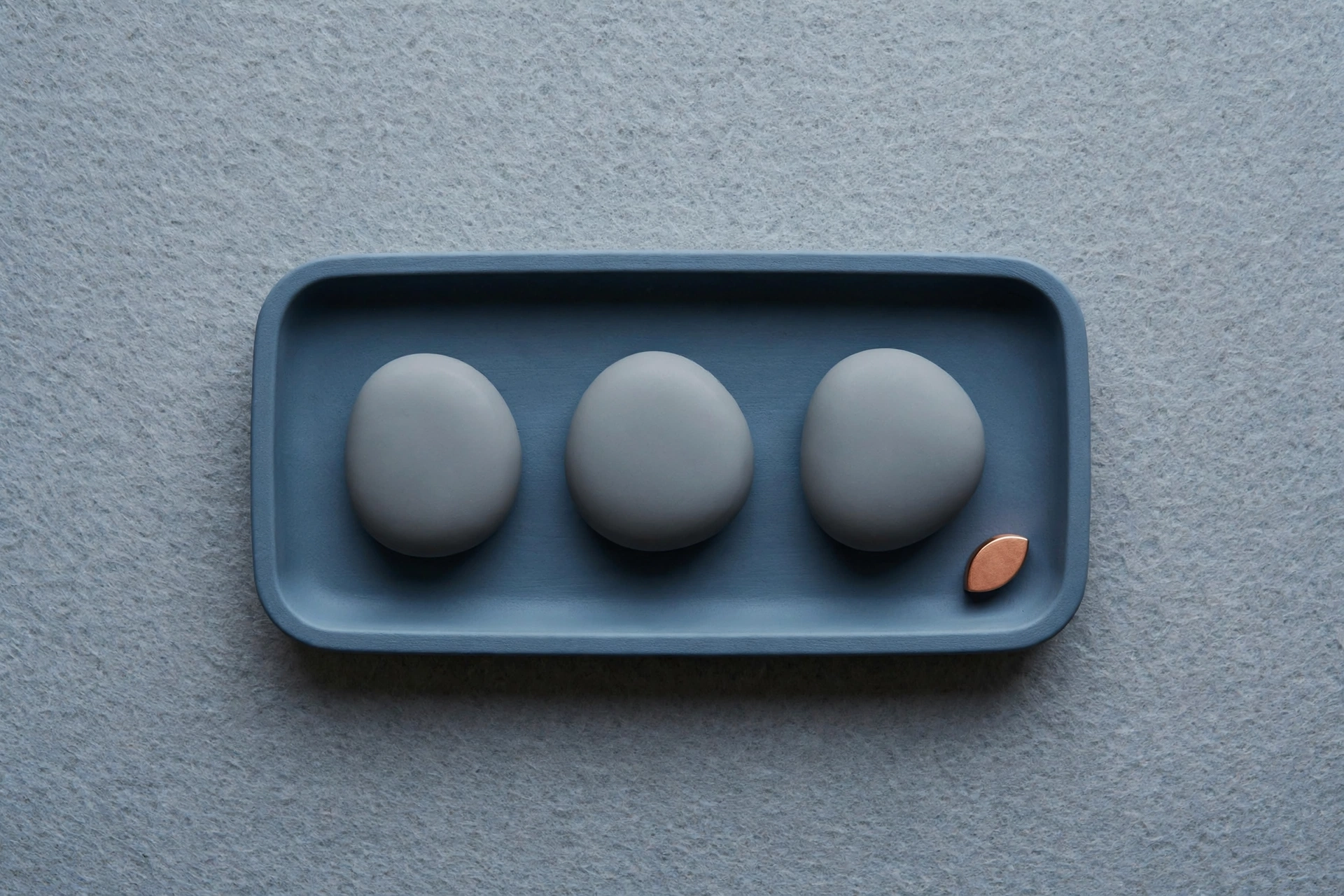 A photograph shows three gray, egg-shaped objects and one copper teardrop-shaped object in a blue rectangular tray on a textured gray surface.