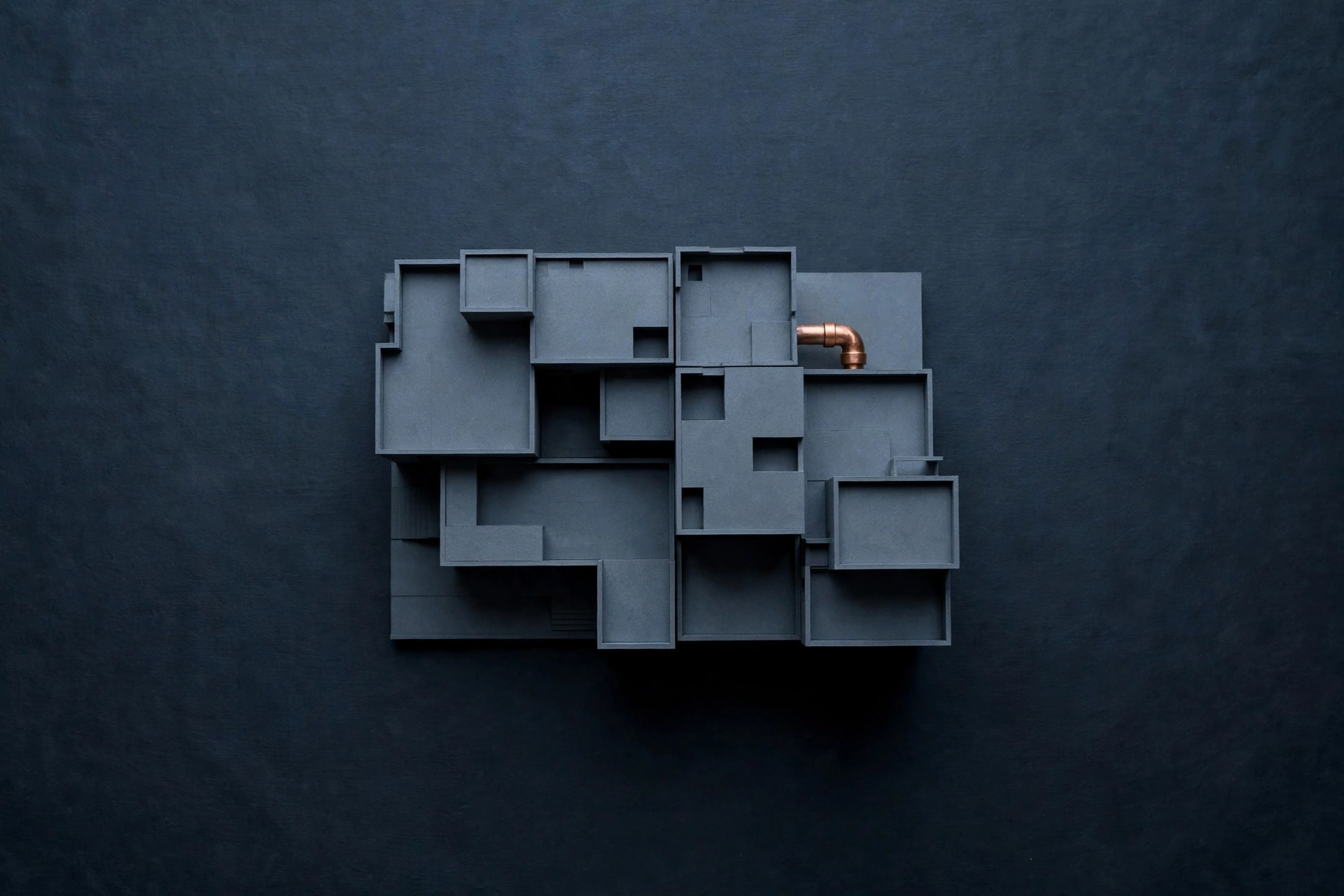 A photograph of a dark grey, multi-level abstract architectural model with a single copper pipe fitting protruding from one corner.