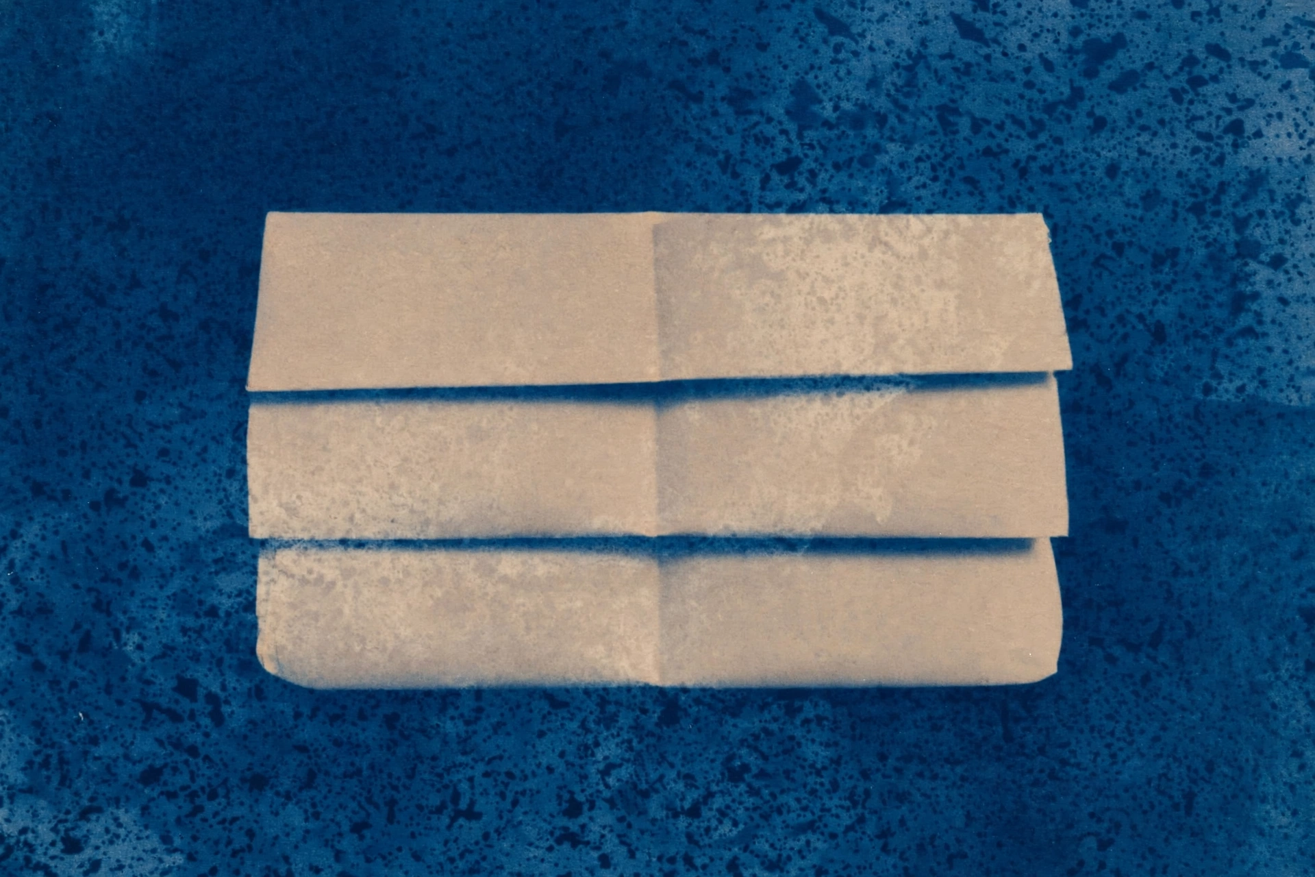 A cyanotype photograph shows folded pieces of beige paper stacked on a dark blue, speckled background.