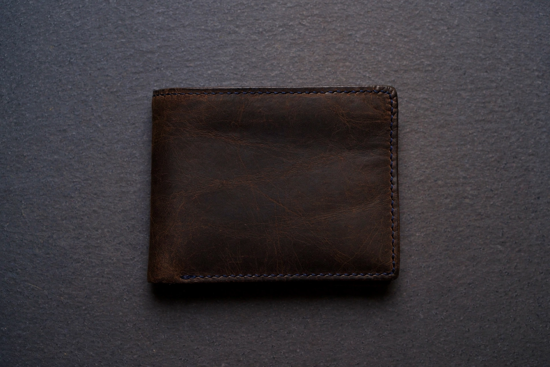 A photograph of a closed brown leather wallet on a dark gray textured surface.