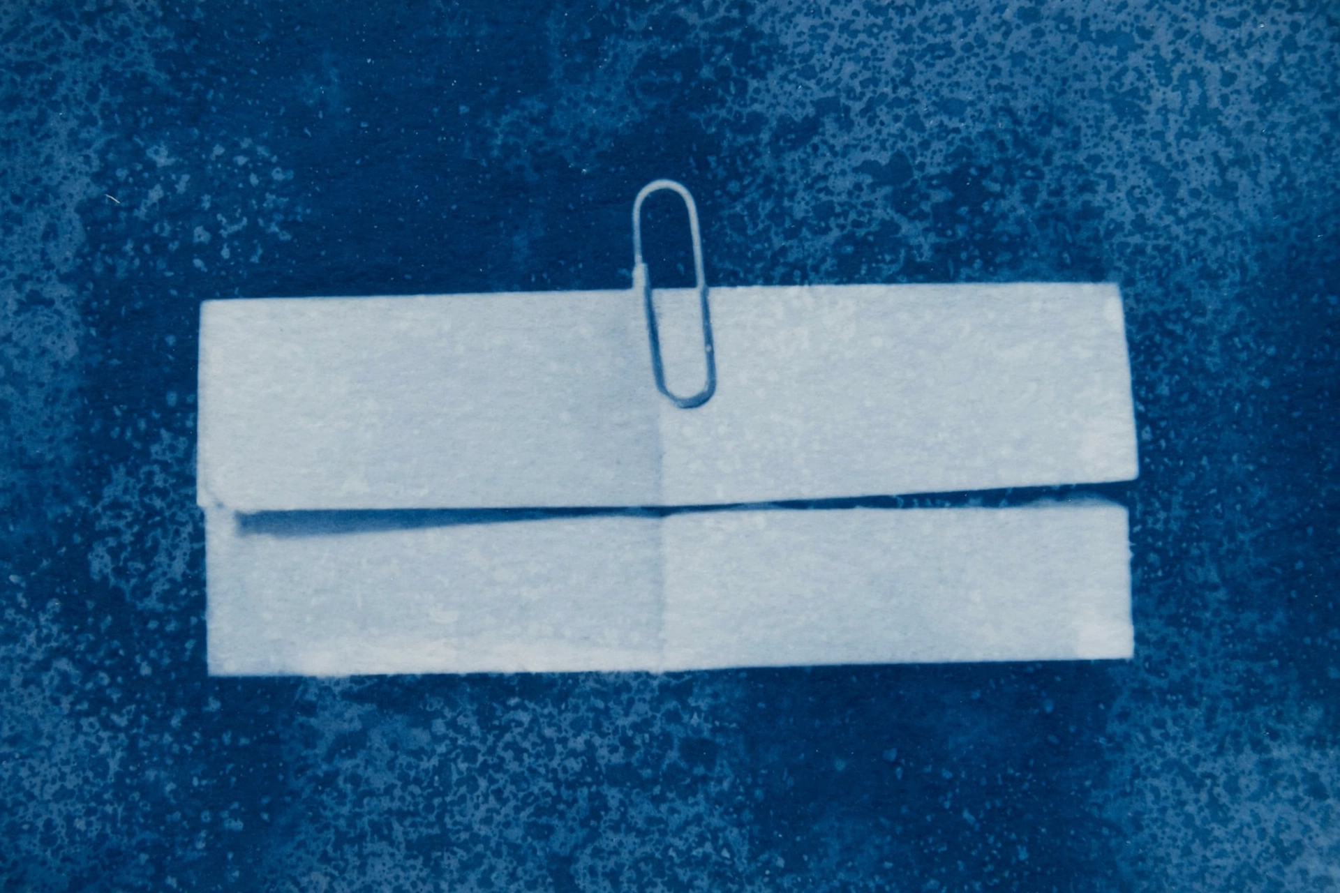 A cyanotype photograph shows a paperclip holding together two pieces of paper.