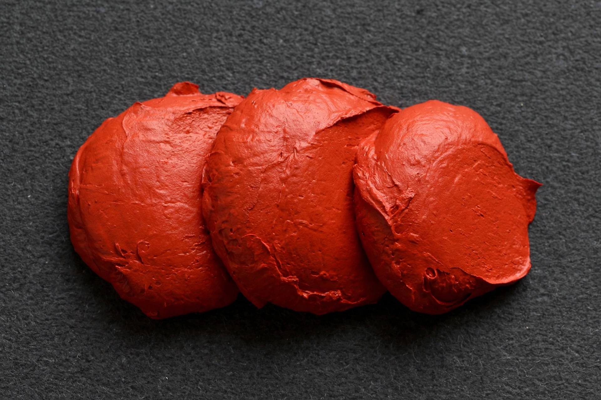 A photograph of three blobs of thick red paint on a textured grey background.