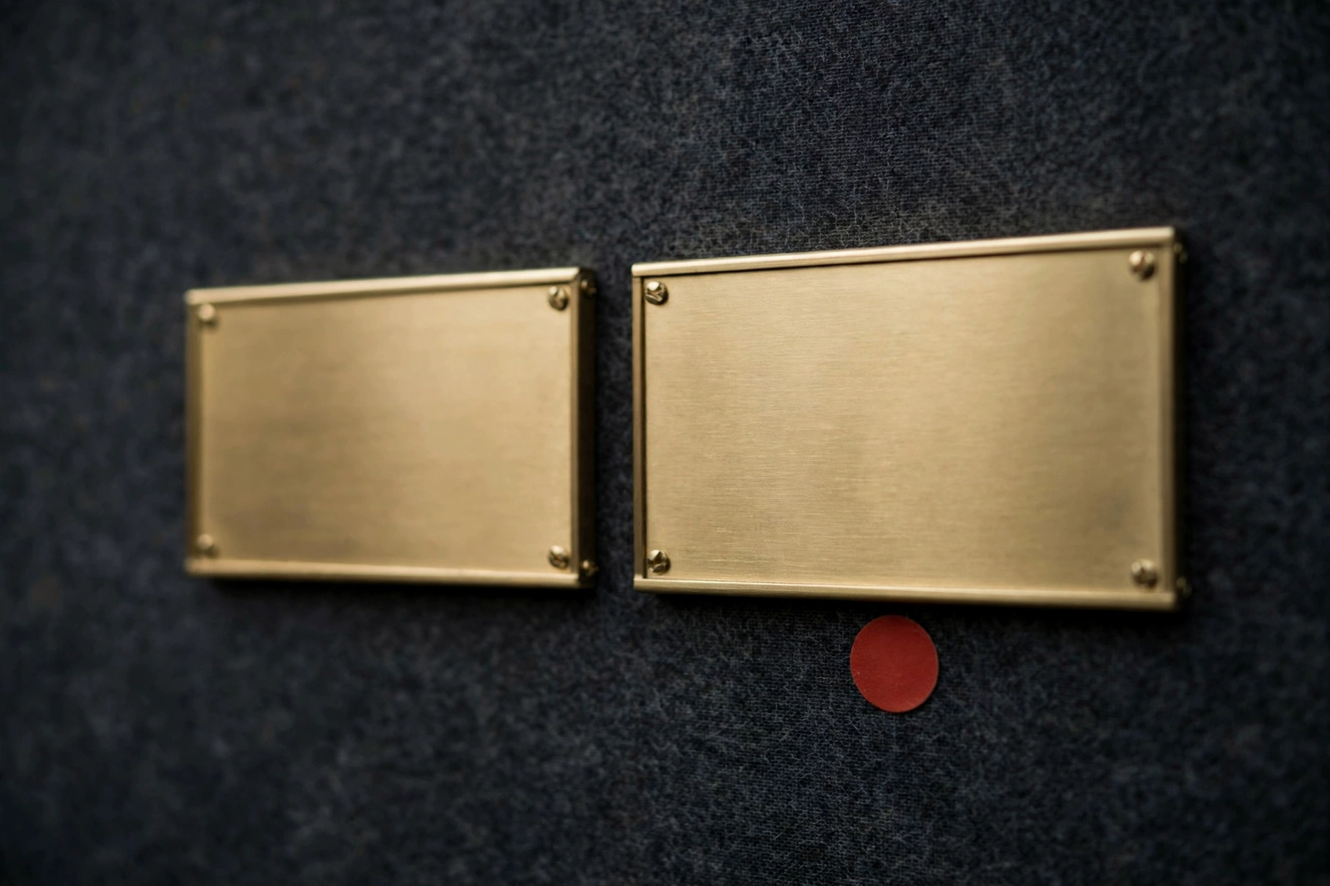 A photograph of two blank brass nameplates affixed to a dark fabric surface, with a small red circle below the right nameplate.
